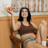 A woman reclining in a wooden chair indoors wearing the Maxomorra GOTS organic cotton women's brief hipster pants in a white, yellow, blue and brown striped design with a brown cotton comfort bra.