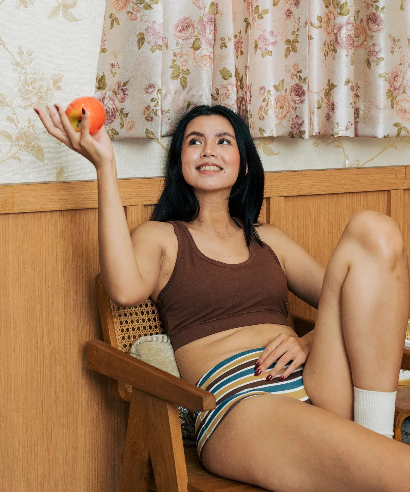 A woman reclining in a wooden chair indoors wearing the Maxomorra GOTS organic cotton women's brief hipster pants in a white, yellow, blue and brown striped design with a brown cotton comfort bra.
