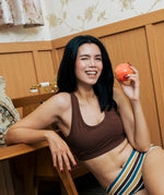 A woman with long black hair smiling while holding a red apple, wearing the Maxomorra GOTS organic cotton women's brief hipster pants in a white, yellow, blue and brown striped design with a brown cotton comfort bra.