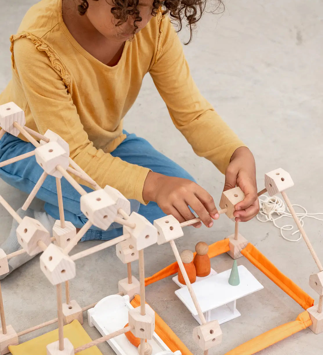 A child building using various wooden poles and blocks from the Trigonos mini large construction set on a concrete surface