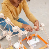 A child building using various wooden poles and blocks from the Trigonos mini large construction set on a concrete surface