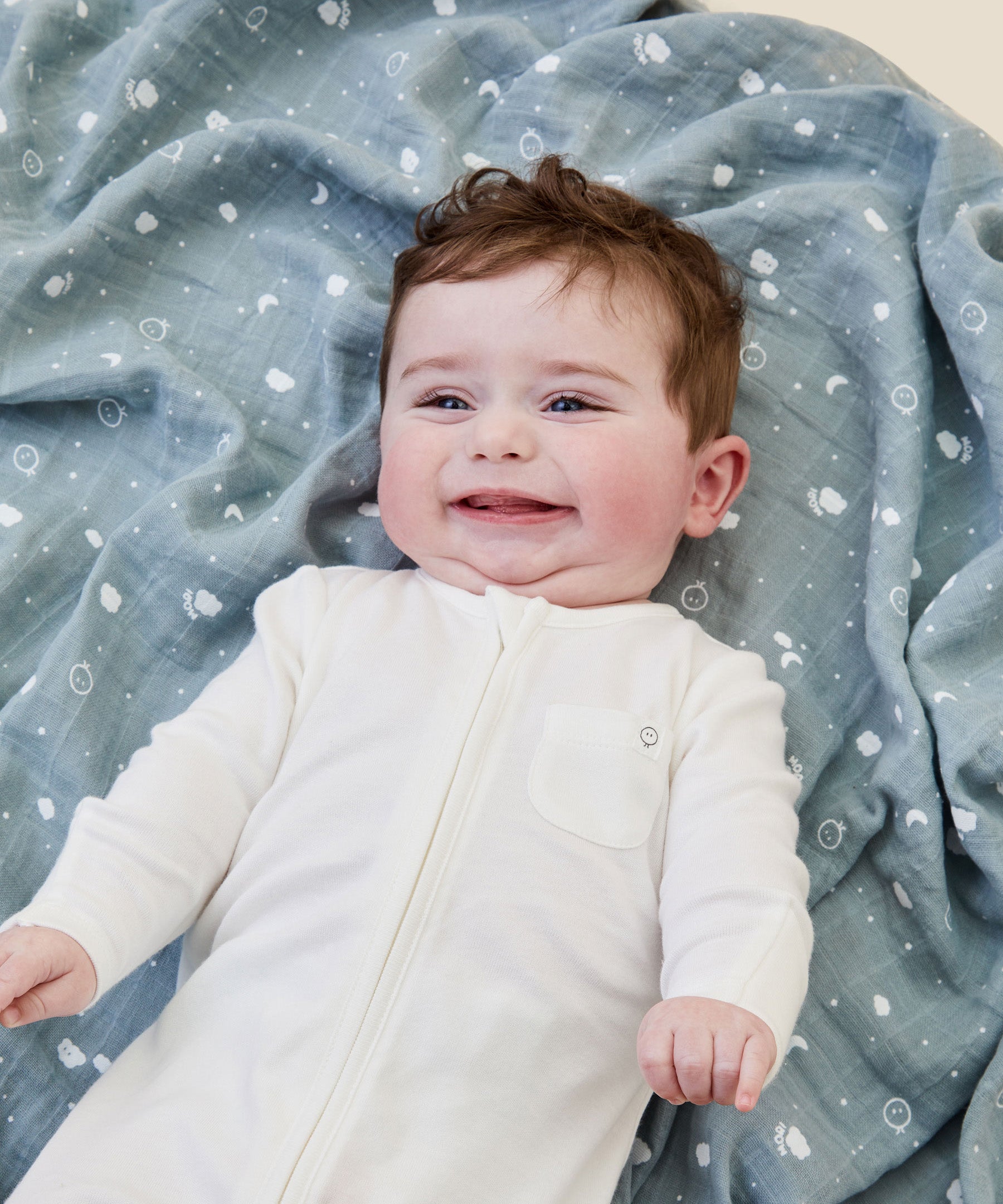A baby lying flat on the blue MORI muslin square showing the cute white prints of clouds, stars and the moon on organic cotton fabric