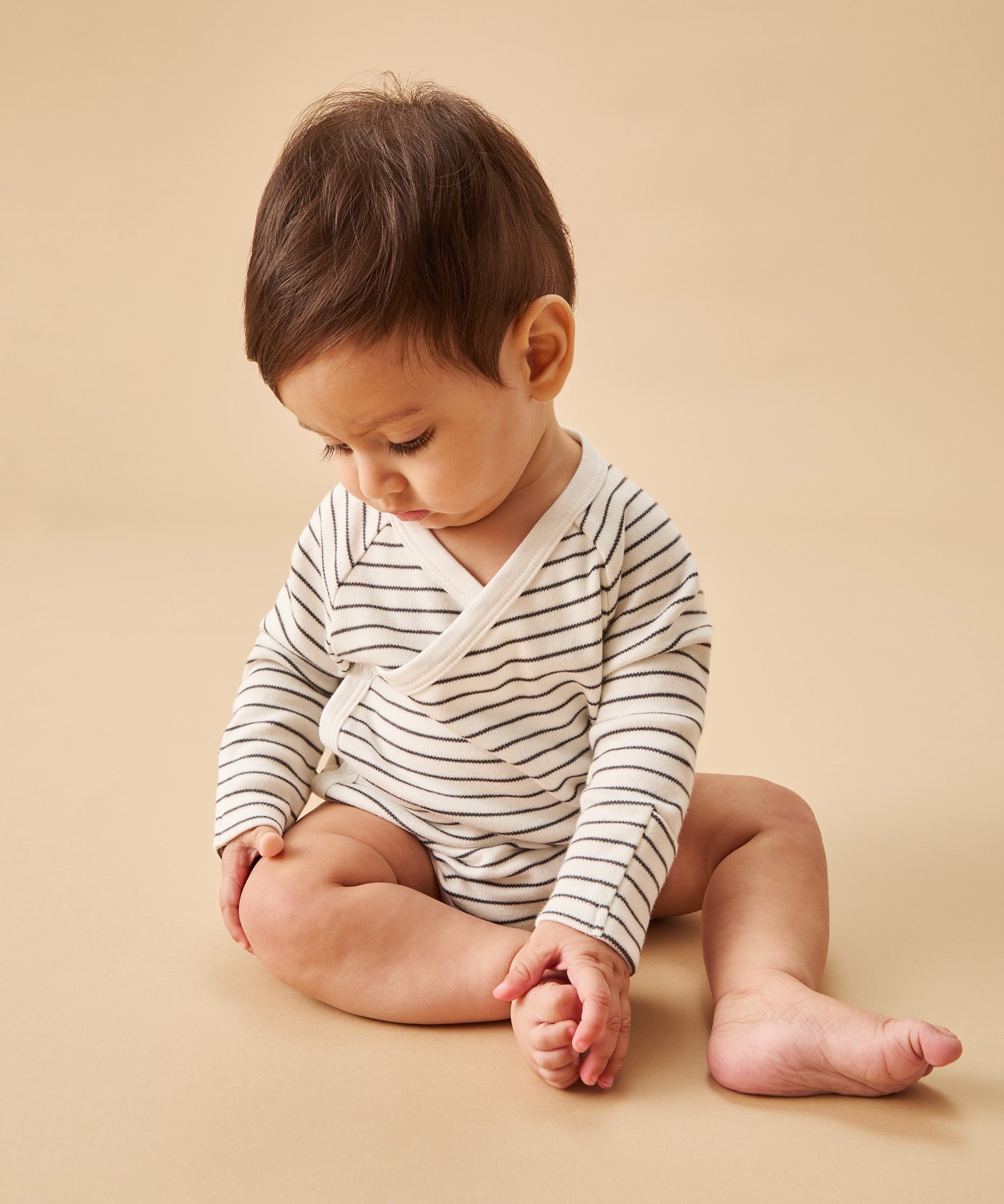 A baby sat upright, wearing a mori organic baby kimono bodysuit, in white with a grey stripy design