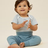 A baby wearing the Mori ribbed comfy joggers in sky blue, and a stripy bodysuit