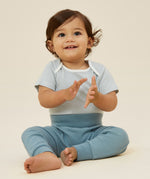 A baby wearing the Mori ribbed comfy joggers in sky blue, and a stripy bodysuit