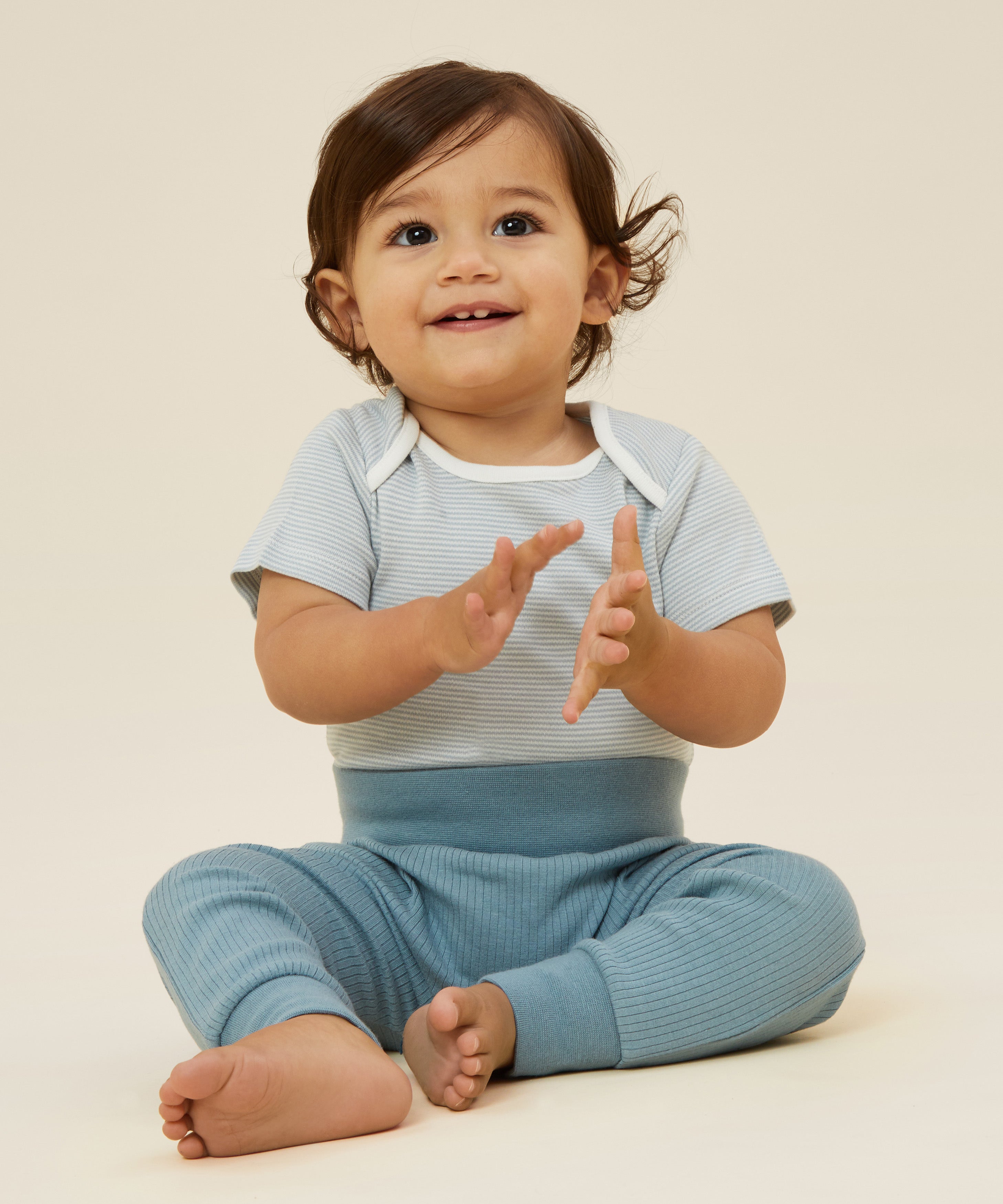 A baby wearing the Mori ribbed comfy joggers in sky blue, and a stripy bodysuit