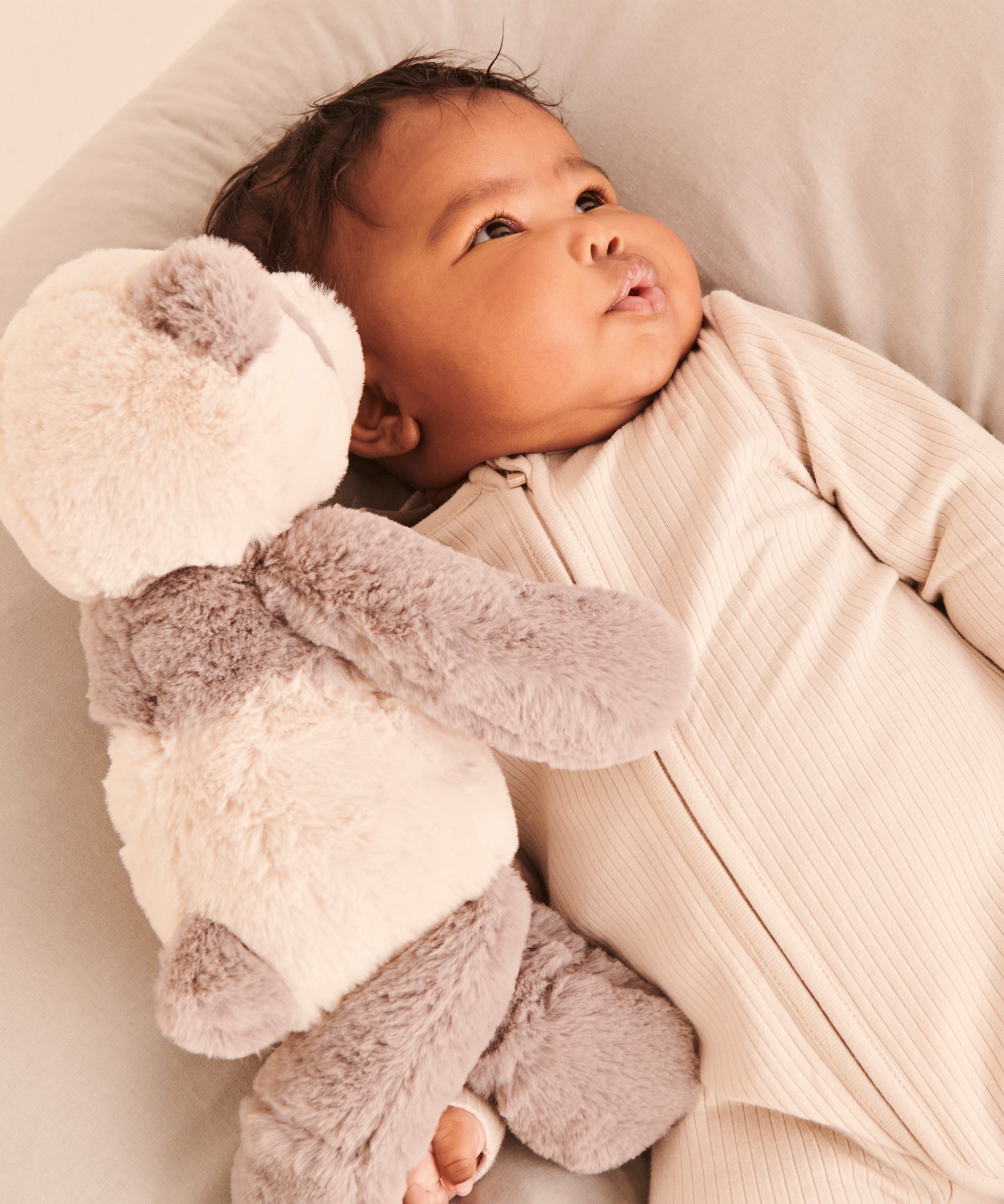 A little baby wearing the Mori clever zipped sleepsuit in Ecru white, and cuddling a teddy bear