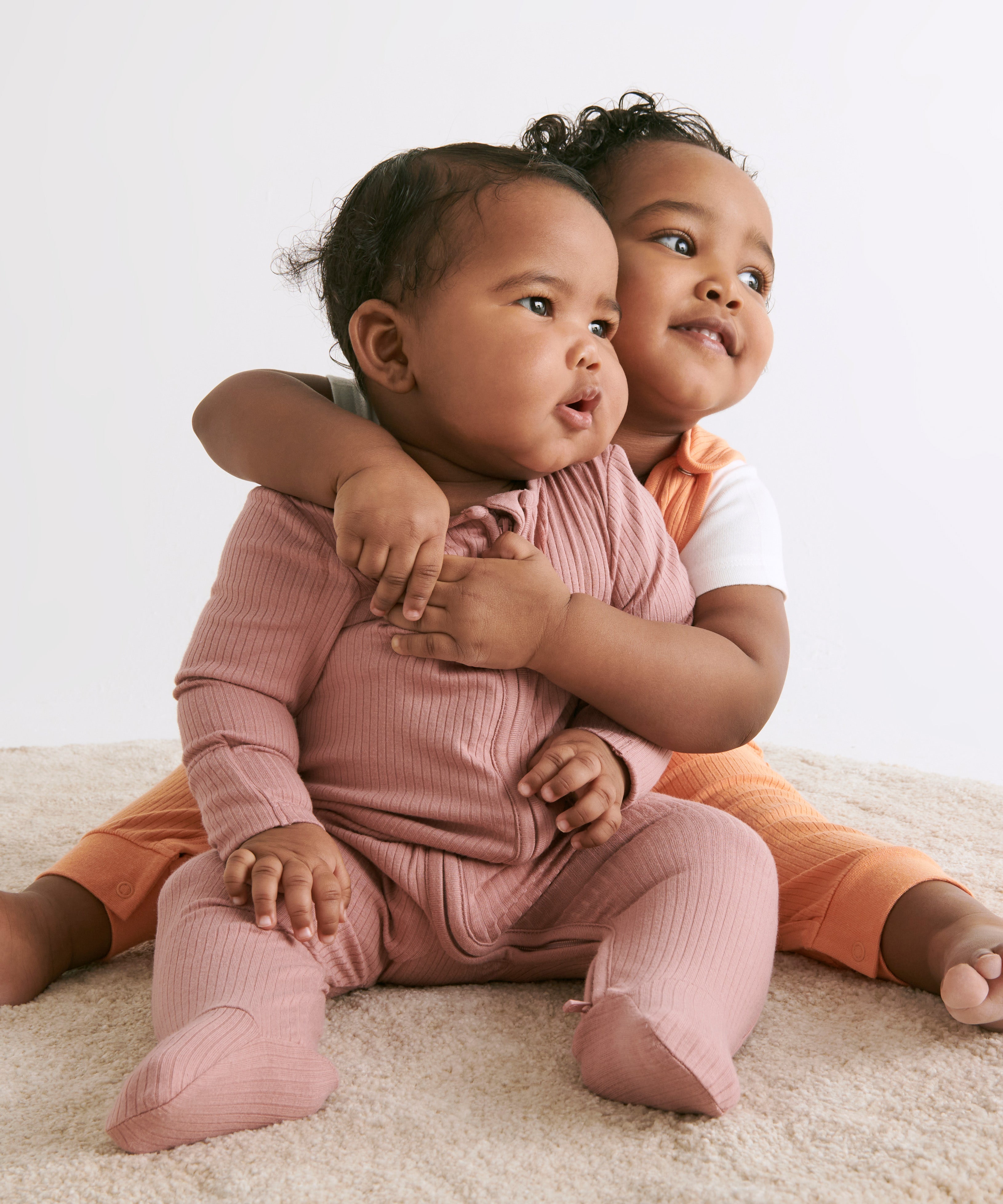 A little baby wearing a rose coloured Mori clever sleepsuit, being hugged by her older toddler sister