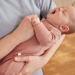 A closeup of a yawning newborn baby being held, wearing a rose coloured Mori clever sleepsuit