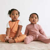 A baby sitting upright with her sister, wearing a rose coloured Mori clever sleepsuit