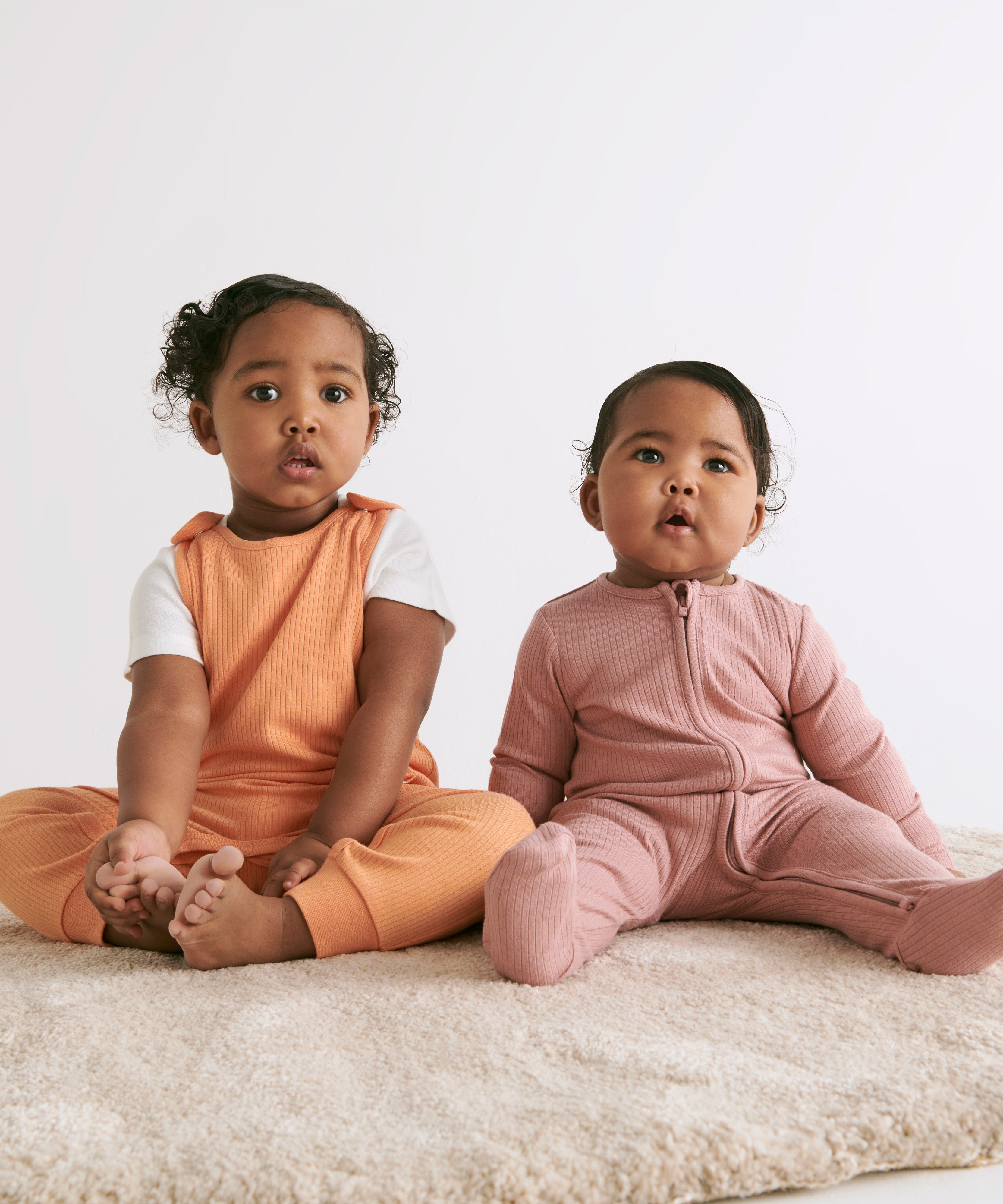 A baby sitting upright with her sister, wearing a rose coloured Mori clever sleepsuit
