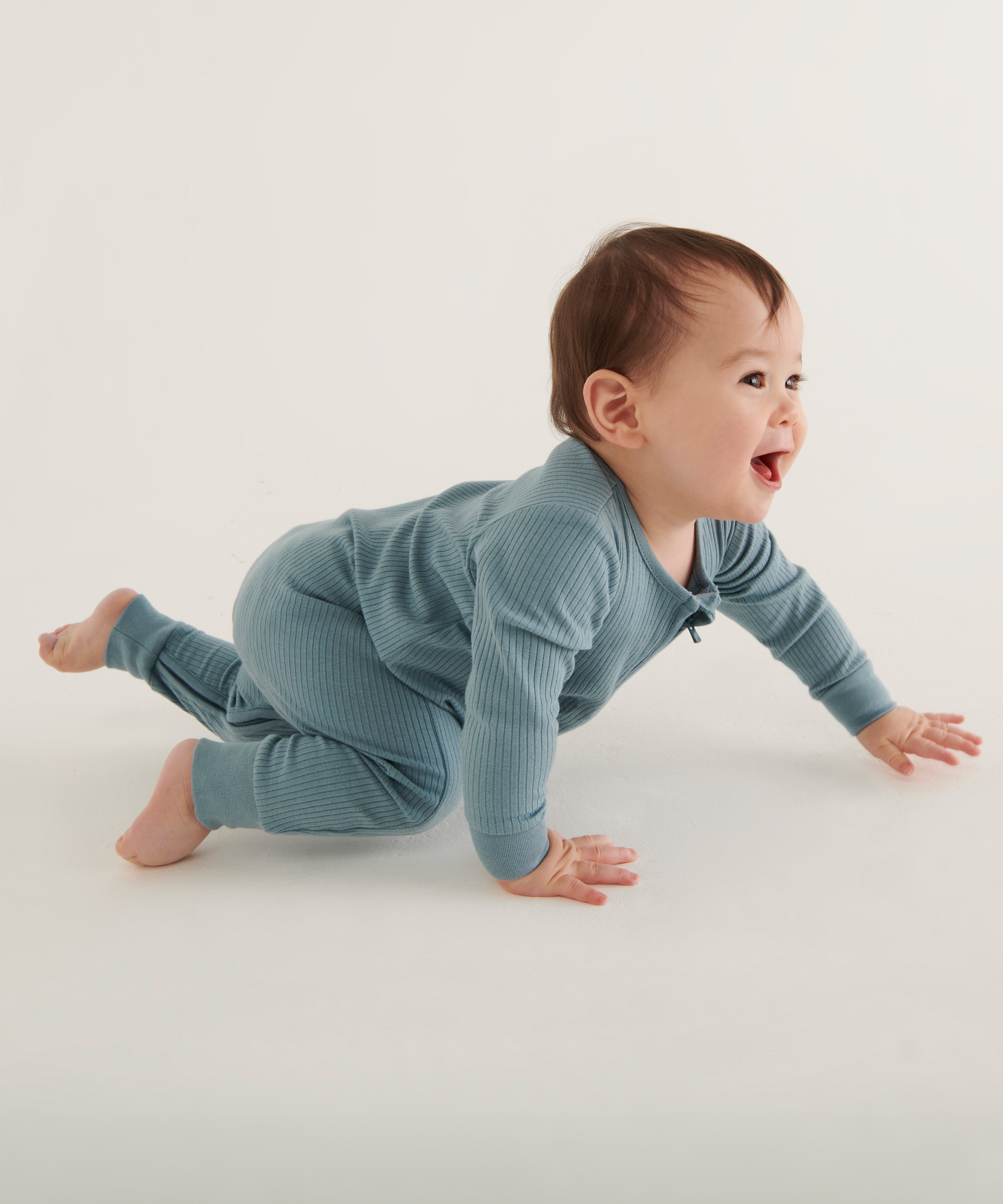 A smiling, crawling, baby wearing the Mori organic zip sleepsuit in a sky blue colour