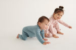 A baby and a toddler wearing Mori organic sleepsuits, one in pink and one in blue