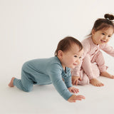 A baby and a toddler wearing Mori organic sleepsuits, one in pink and one in blue