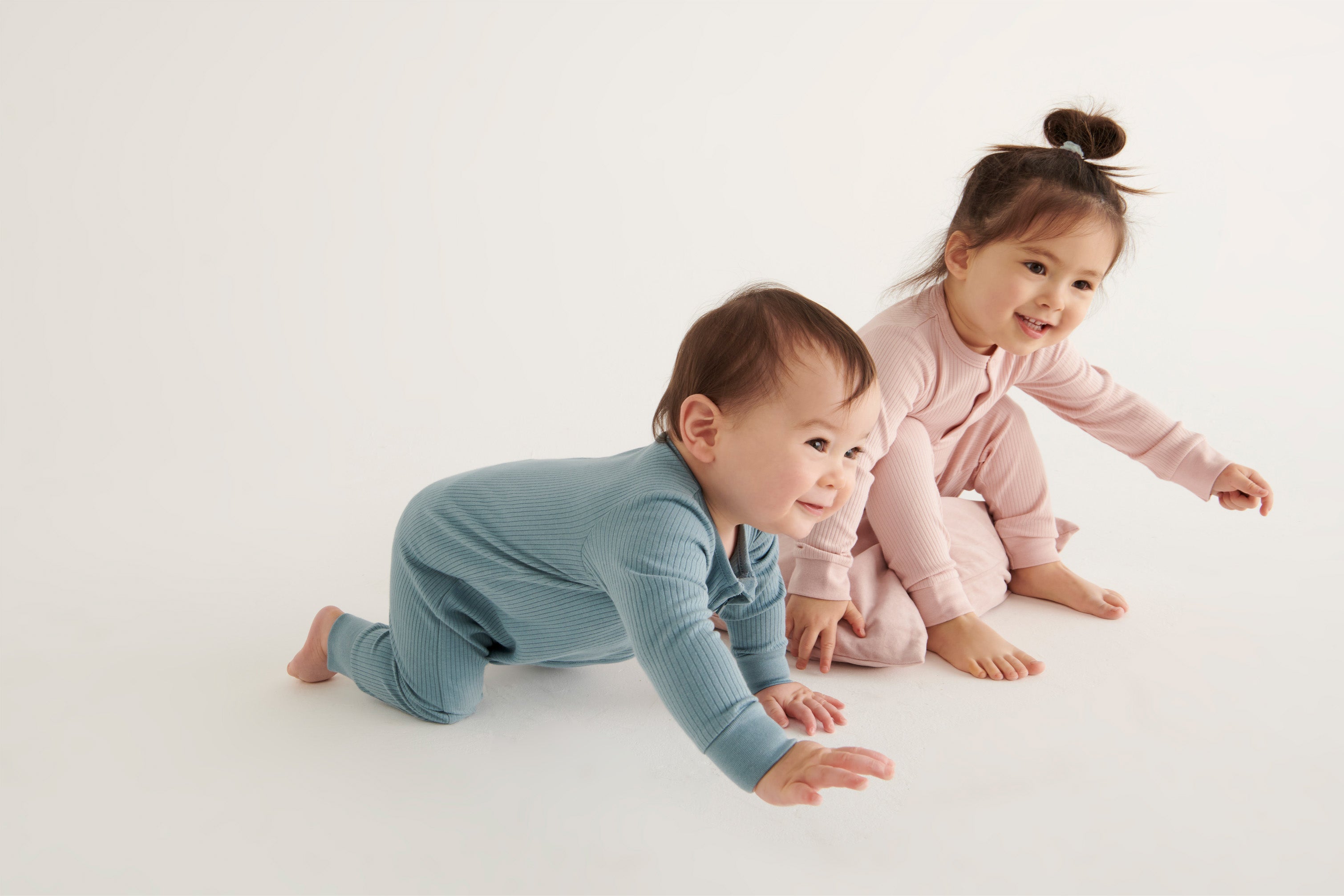 A baby and a toddler wearing Mori organic sleepsuits, one in pink and one in blue