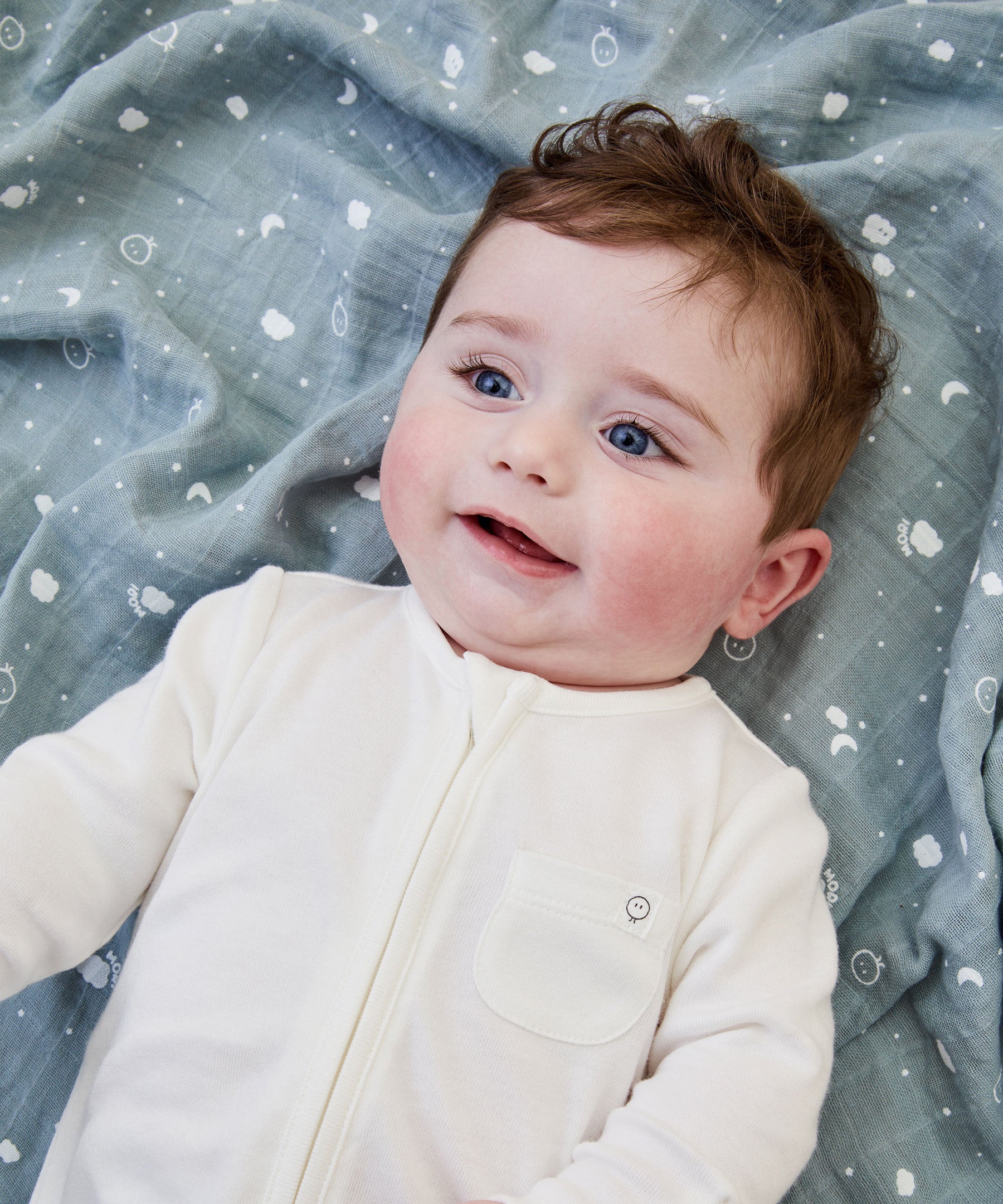 A close up of a baby lying flat on the blue MORI muslin square showing the cute white prints of clouds, stars and the moon on soft organic cotton fabric