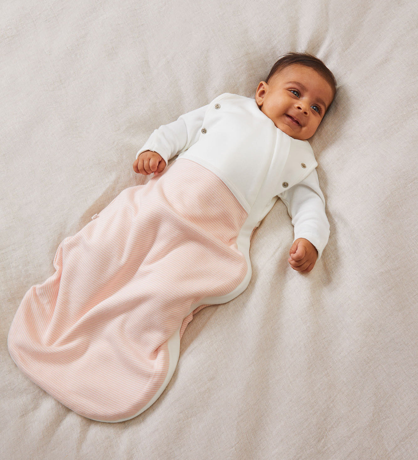 A baby laid on a soft surface inside the blush stripe MORI swaddle. Made from bamboo and organic cotton and features poppers on the arms, to tuck the arms in or let them be free.