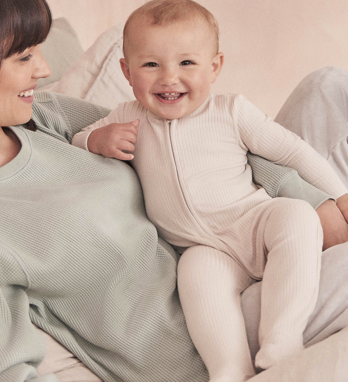 A baby wearing the Mori two way zipped sleepsuit in ecru colour. A ribbed style all in one outfit made from bamboo and organic cotton. Features a two-way zip and concealed for safety