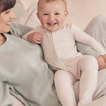 A baby wearing the Mori two way zipped sleepsuit in ecru colour. A ribbed style all in one outfit made from bamboo and organic cotton. Features a two-way zip and concealed for safety