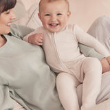 A baby wearing the Mori two way zipped sleepsuit in ecru colour. A ribbed style all in one outfit made from bamboo and organic cotton. Features a two-way zip and concealed for safety