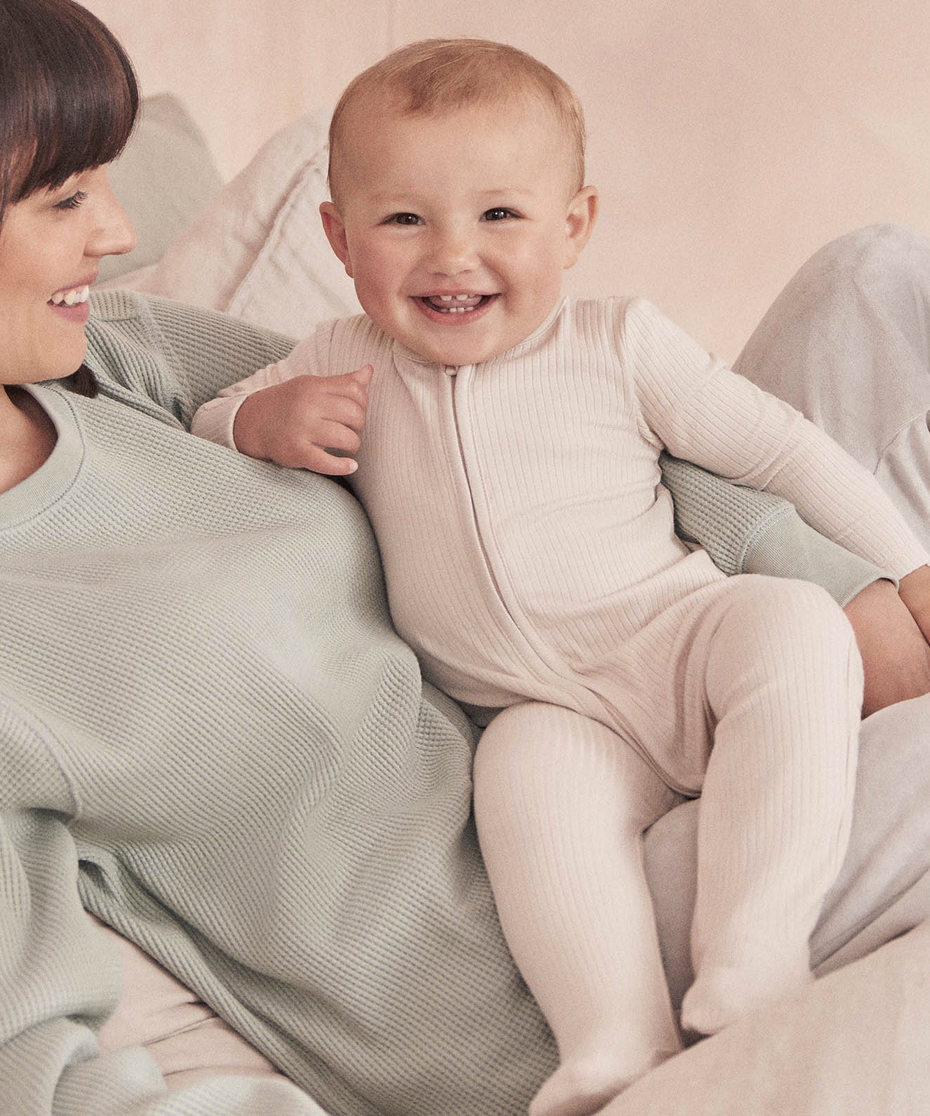 A baby wearing the Mori two way zipped sleepsuit in ecru colour. A ribbed style all in one outfit made from bamboo and organic cotton. Features a two-way zip and concealed for safety