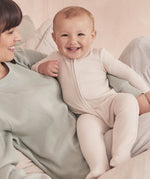 A baby wearing the Mori two way zipped sleepsuit in ecru colour. A ribbed style all in one outfit made from bamboo and organic cotton. Features a two-way zip and concealed for safety