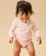 A toddler standing up, and wearing the Mori organic cotton and bamboo blend baby kimono bodysuit in a pink blush stripe design.