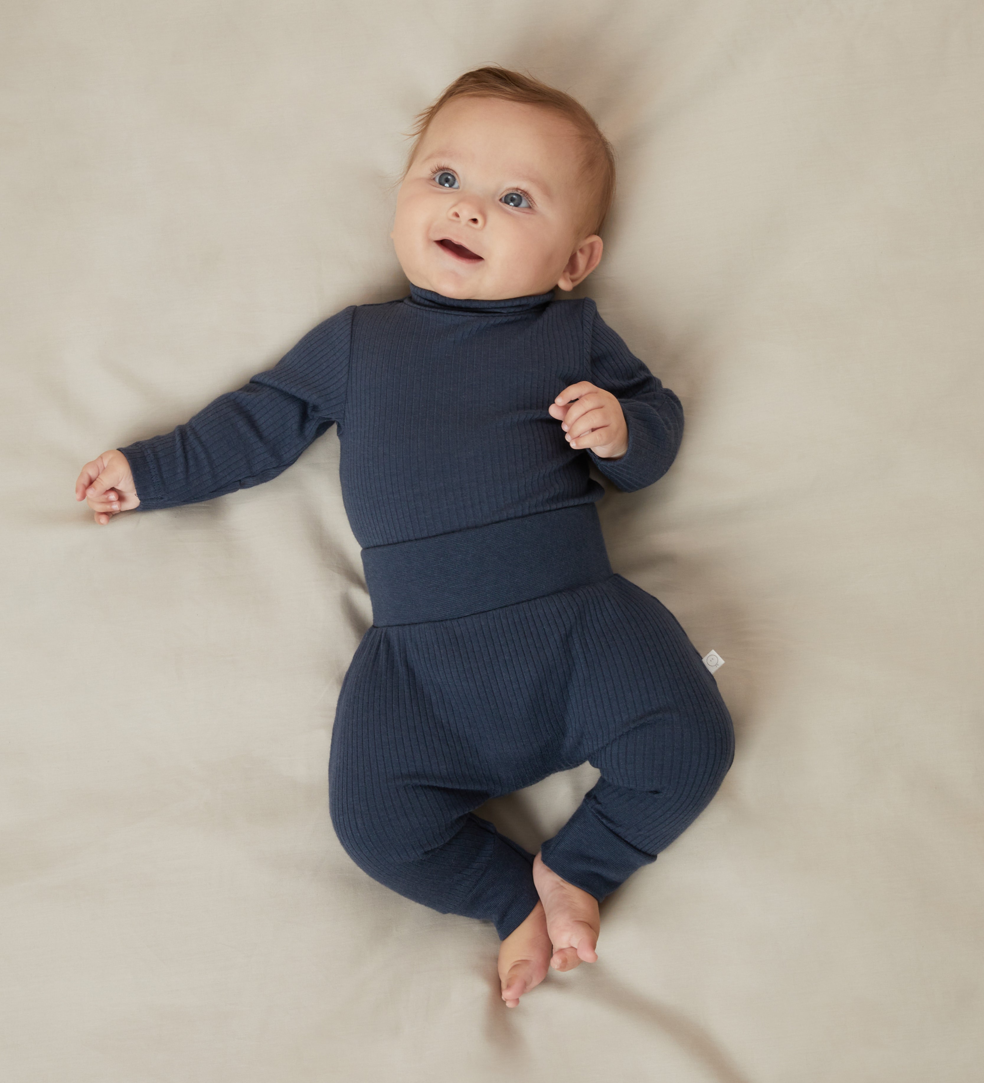 A baby laying on a bed, wearing a matching set of Mori navy comfy joggers made from organic cotton, and a long sleeve top.