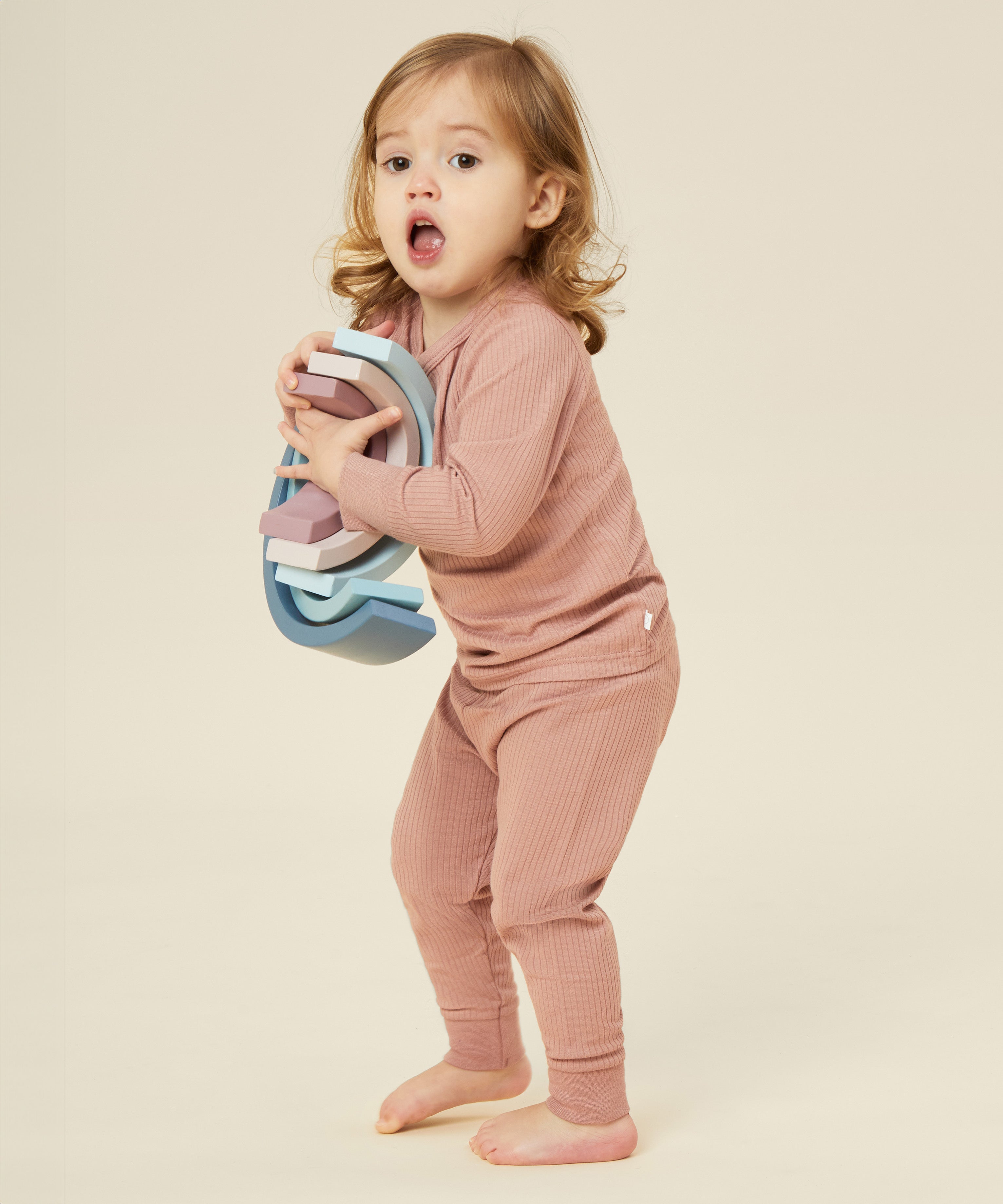 A little girl holding a wooden rainbow stacker toy, and wearing the Mori organic cotton and bamboo blend pyjamas in pink.