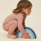 A little girl playing with a wooden rainbow stacker toy, and wearing the Mori organic cotton and bamboo blend pyjamas in pink.