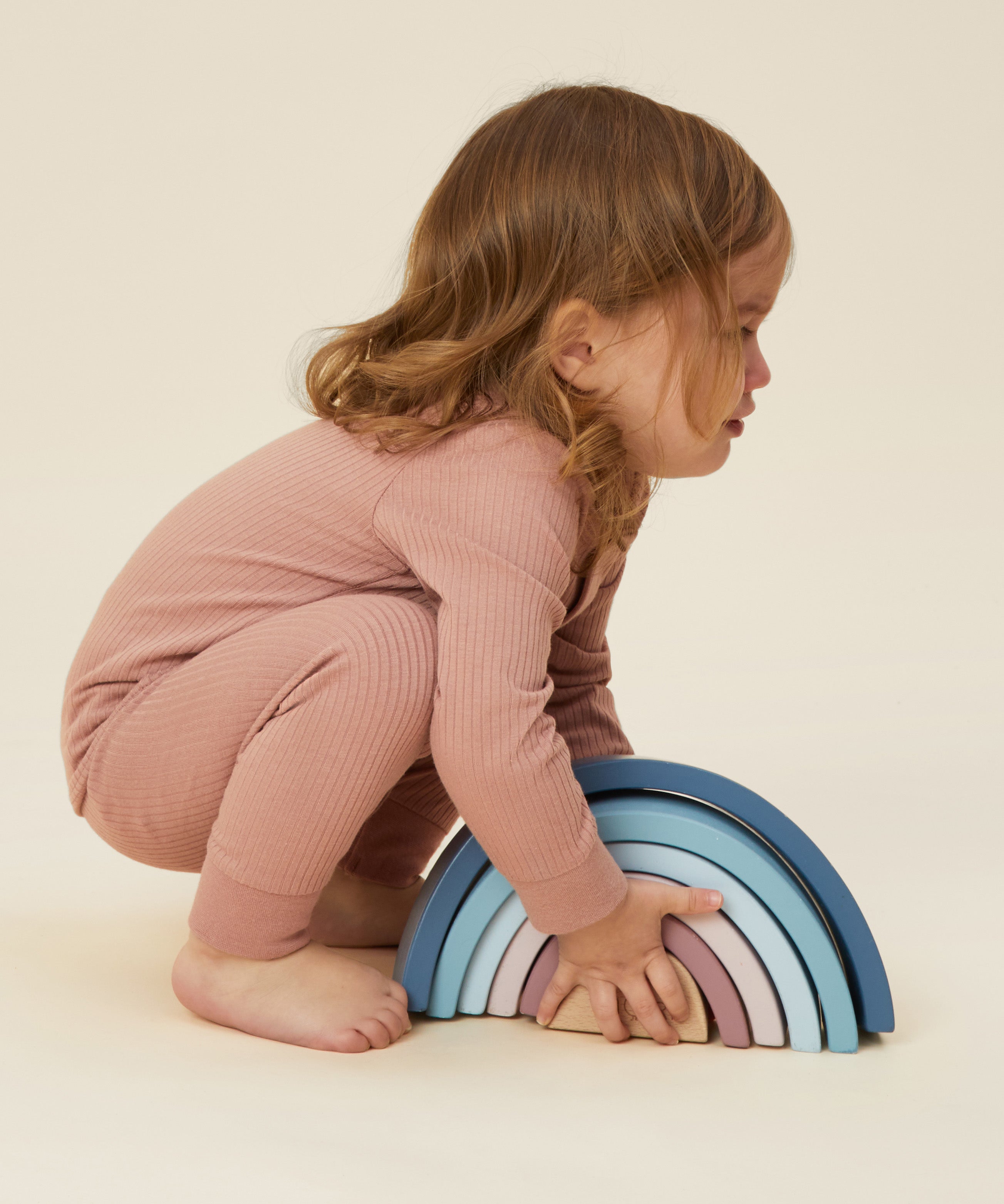 A little girl playing with a wooden rainbow stacker toy, and wearing the Mori organic cotton and bamboo blend pyjamas in pink.