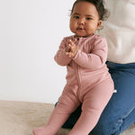 A baby sitting on the mother's knees wearing the MORI two way zip ribbed sleepsuit in rose colour.