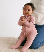 A baby sitting on the mother's knees wearing the MORI two way zip ribbed sleepsuit in rose colour.