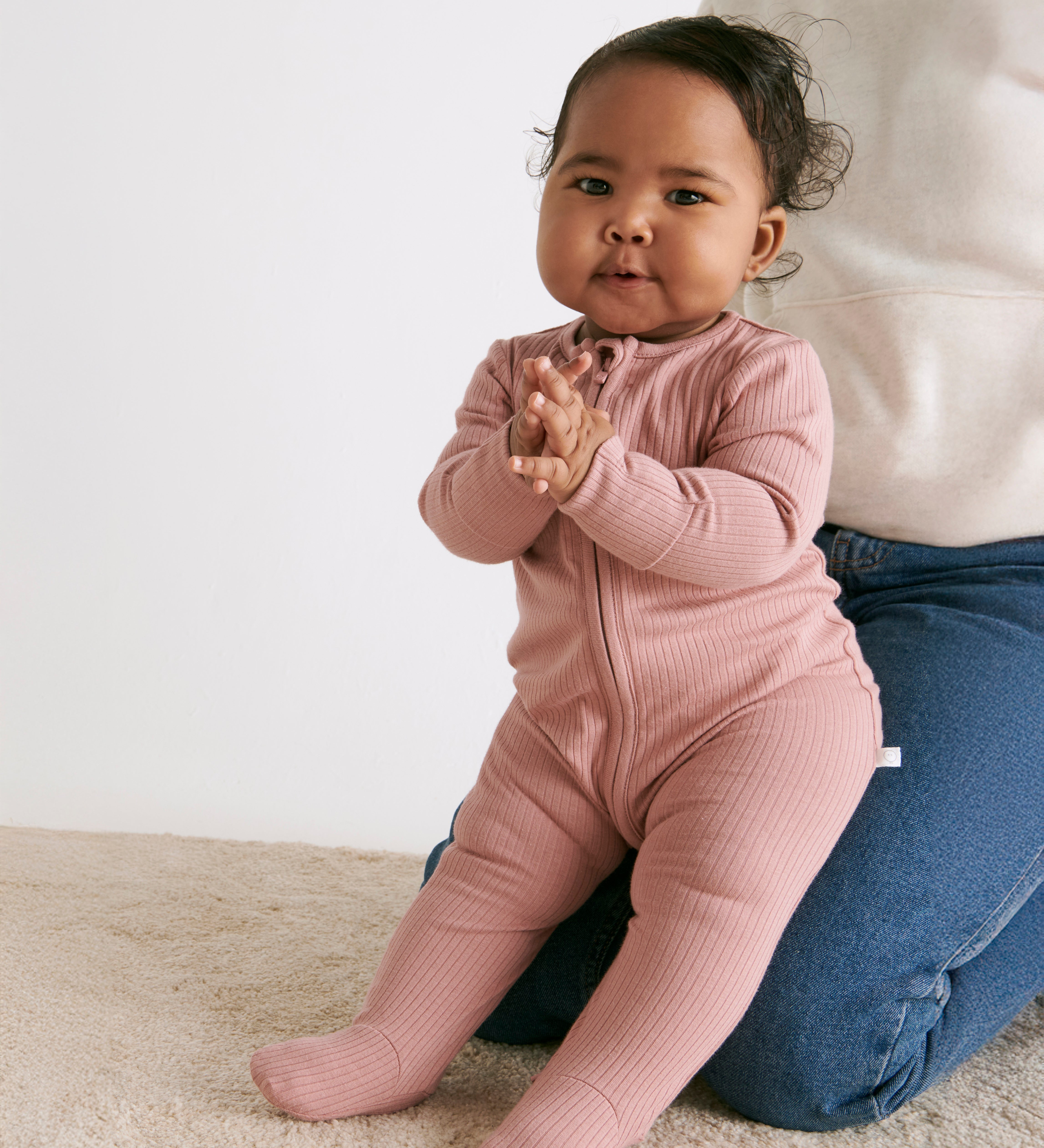 A baby sitting on the mother's knees wearing the MORI two way zip ribbed sleepsuit in rose colour.