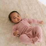 A baby laying flat on a fluffy rug on the mother's knees wearing the MORI two way zip ribbed sleepsuit in rose colour.