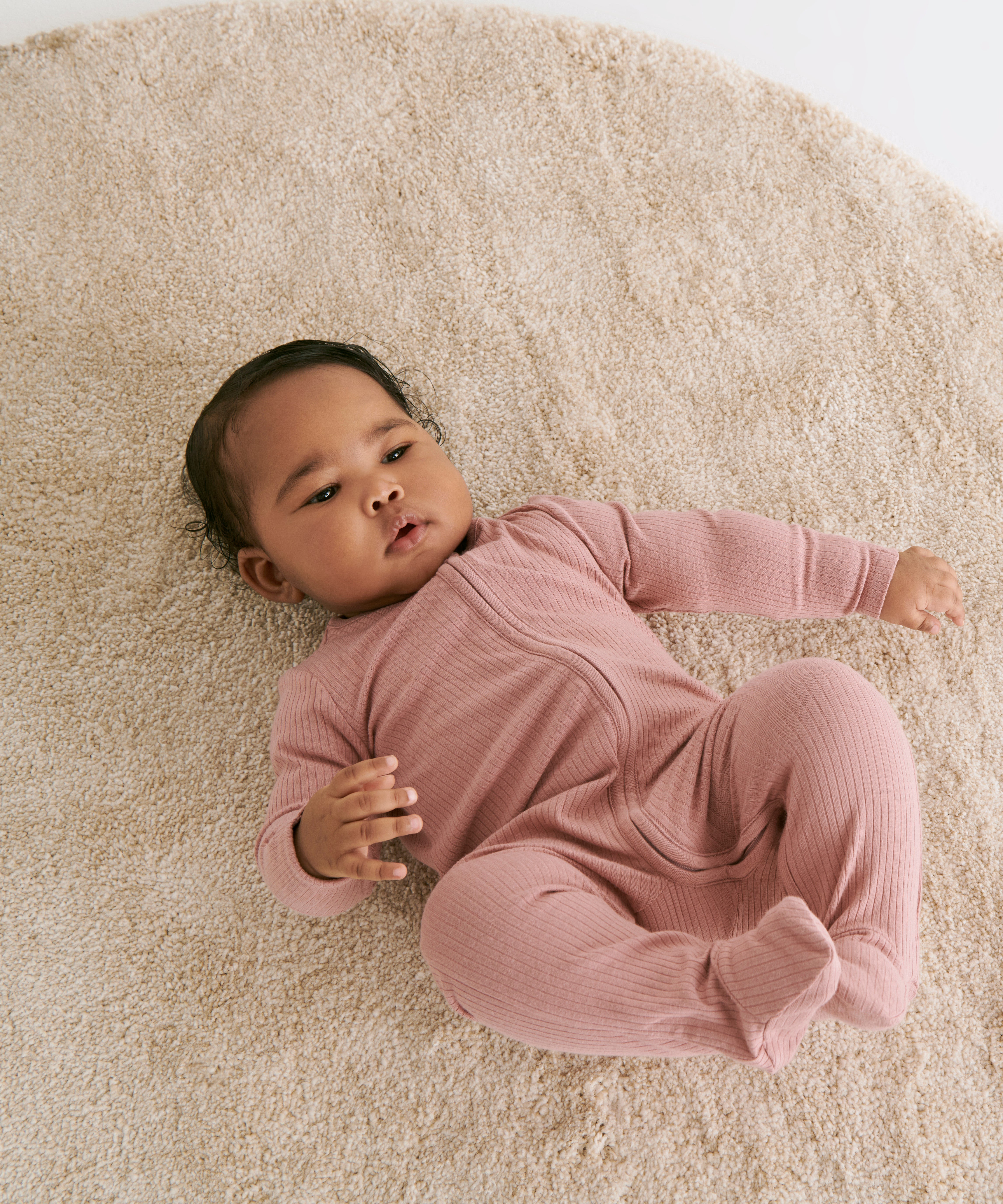 A baby laying flat on a fluffy rug on the mother's knees wearing the MORI two way zip ribbed sleepsuit in rose colour.