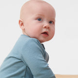 A close up of a  baby sitting  wearing the MORI two way zip ribbed sleepsuit in sky blue colour.
