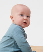 A close up of a  baby sitting  wearing the MORI two way zip ribbed sleepsuit in sky blue colour.