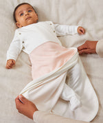 A baby laid on a soft surface inside the blush stripe MORI swaddle. Showing adults hand zipping the bottom half of the swaddle to tuck the baby in.