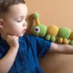 Boy stood in front of a beige wall with the Myum handmade crochet caterpillar toy balanced on his arm