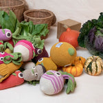 Myum handmade soft crochet vegetable toys scattered in a pile on a white table next to come real vegetables and wooden baskets