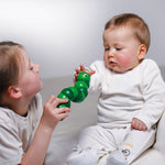 Young girl playing with a green Naef Juba worm toy in front of a baby who is reaching out to hold it.