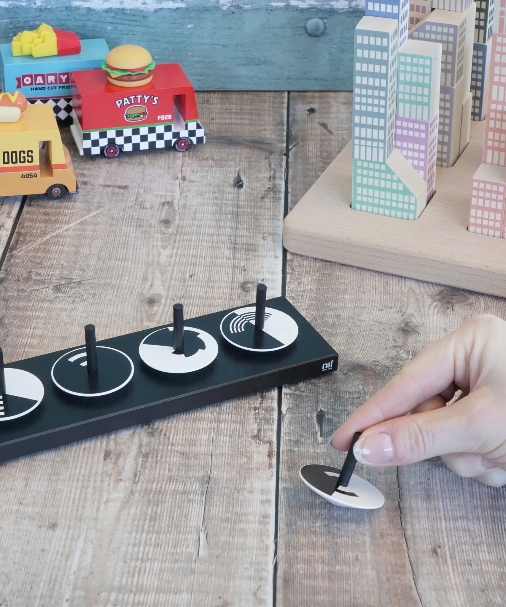 Adult hand holding a single Naef wooden optical illusion spinning top on a wooden surface with vehicles and building blocks in the background