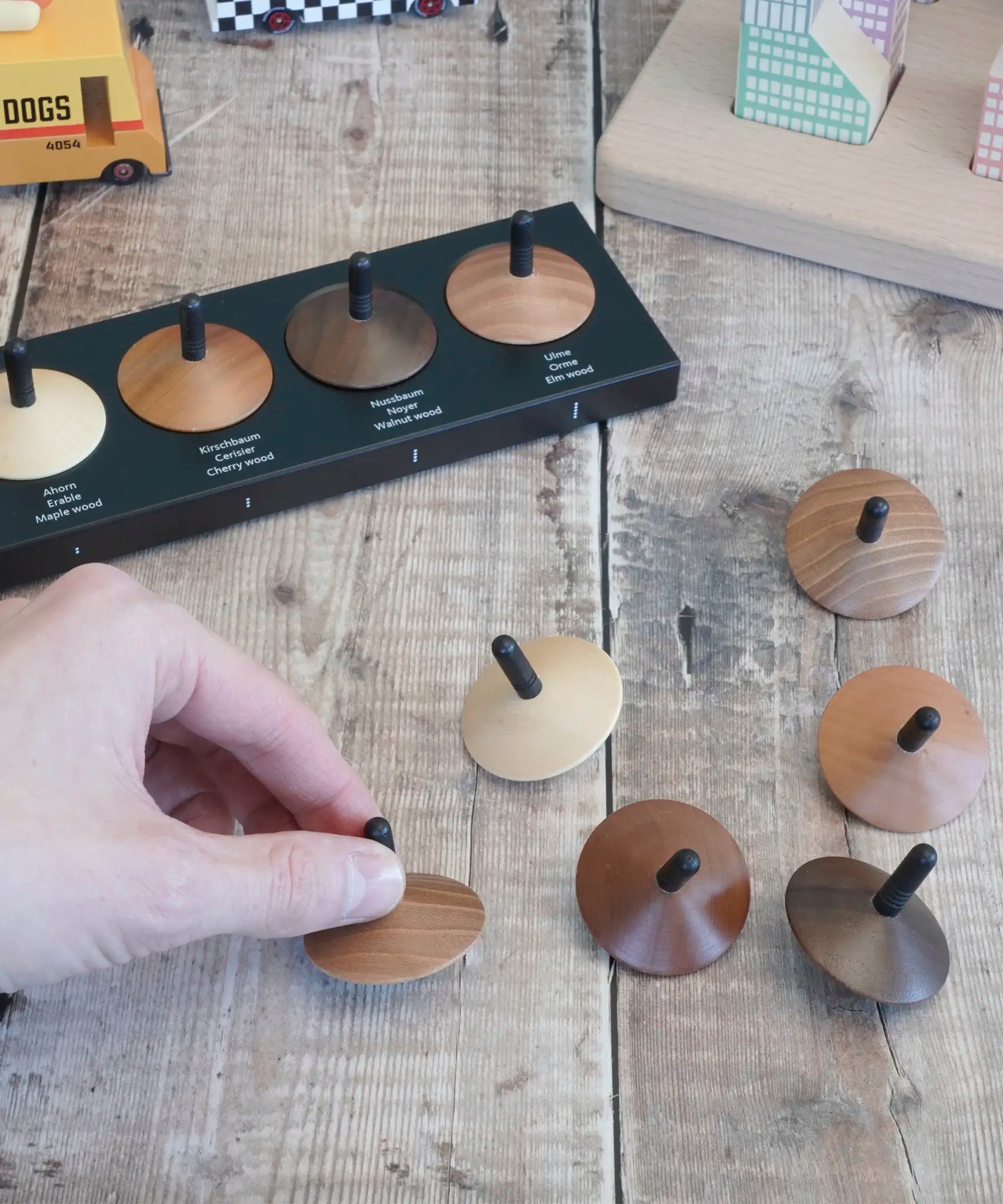 Adult hand holding a loose Naef wooden spinning top by the black stem on a wooden surface next to other spinning tops