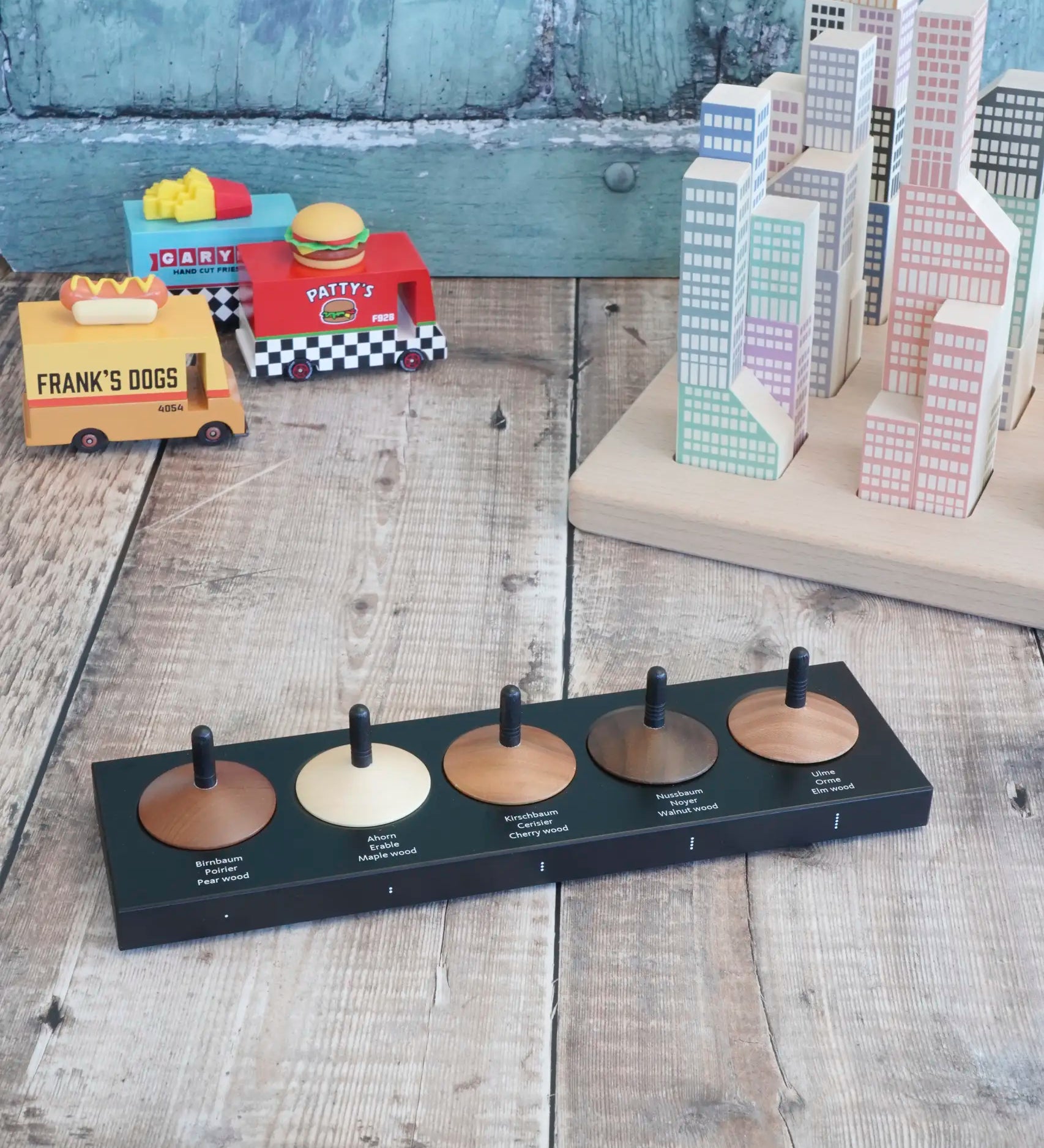 Naef wooden spinning top set sitting on the black holder showing names of different wood with vehicles and blocks in the background.