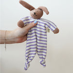 A Nanchen Rabbit Baby Comforter being held up in an adult's hand. The main body of the comforter is made of a white and lilac purple coloured striped, ribbed material.