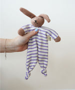 A Nanchen Rabbit Baby Comforter being held up in an adult's hand. The main body of the comforter is made of a white and lilac purple coloured striped, ribbed material.