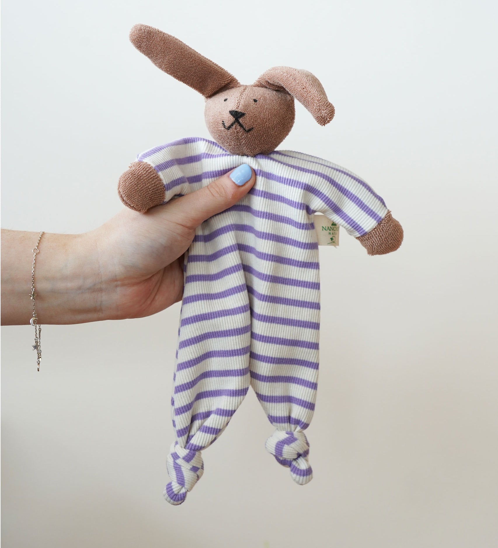 A Nanchen Rabbit Baby Comforter being held up in an adult's hand. The main body of the comforter is made of a white and lilac purple coloured striped, ribbed material.