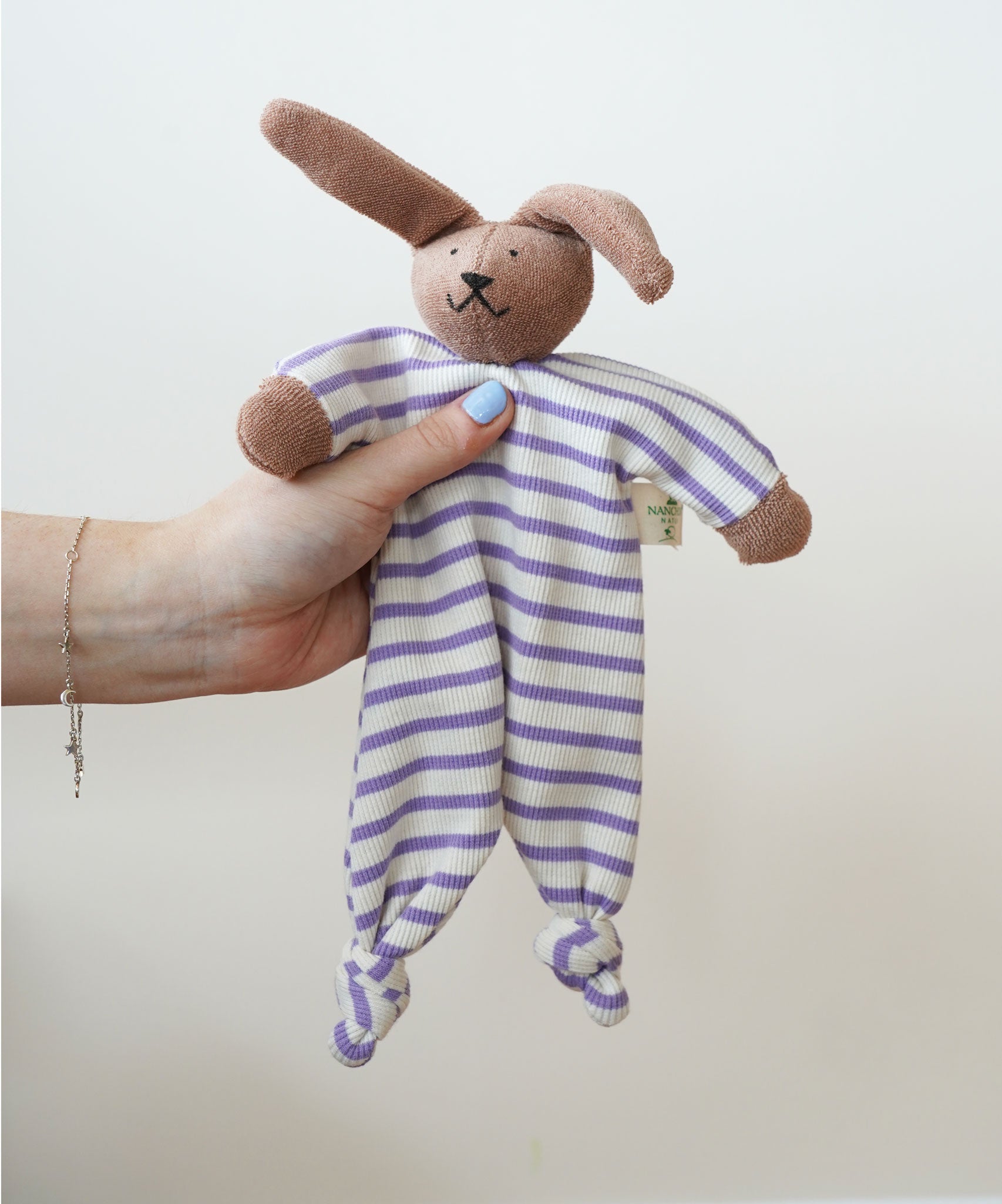 A Nanchen Rabbit Baby Comforter being held up in an adult's hand. The main body of the comforter is made of a white and lilac purple coloured striped, ribbed material.