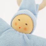 Close-up of the Nanchen Pimpel Doll Baby Comforter in soft blue. The doll's face features gently painted eyes, cheeks, and lips, framed by a light blue organic cotton terry hood. A wooden teething ring is partially visible behind the pointed hat.